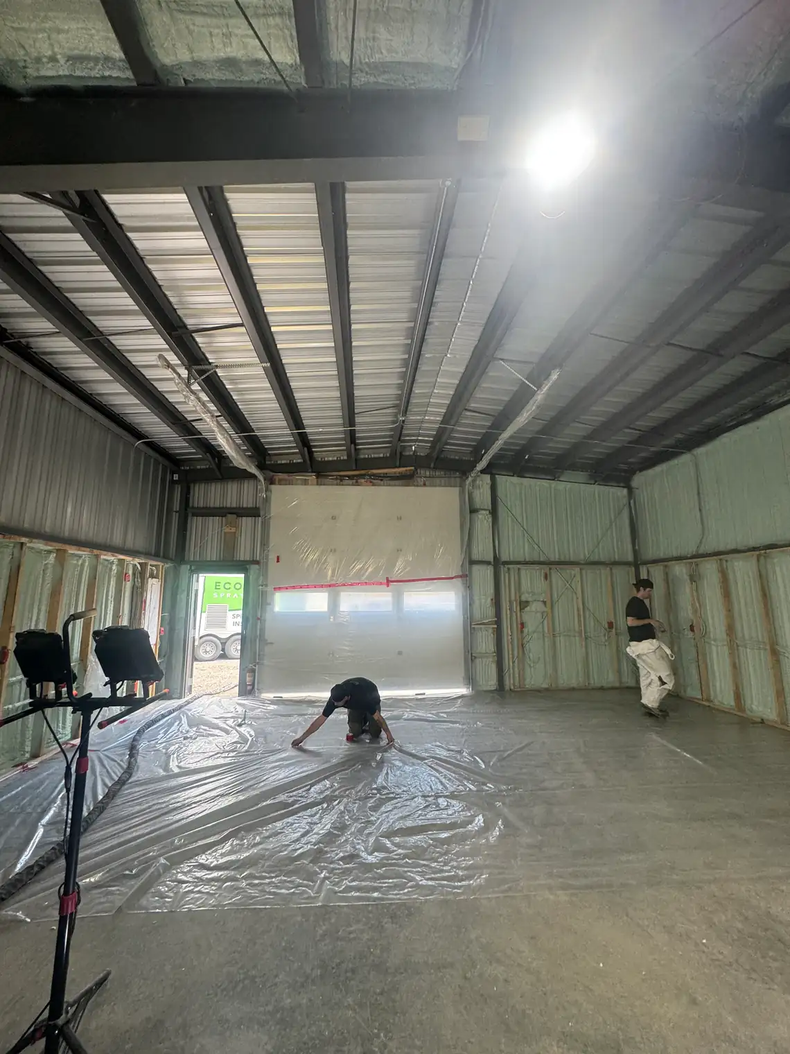 Two EcoSeal crew members taping plastic sheeting across a shop floor, with the walls already sprayed and the steel roof deck still bare above