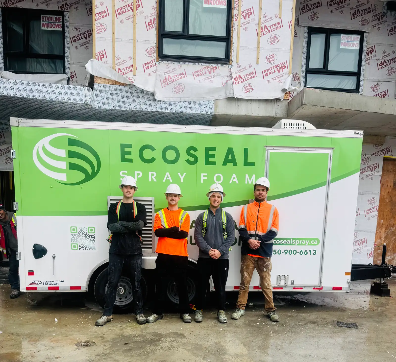 Four-person EcoSeal crew in hard hats in front of the EcoSeal spray foam trailer at a Protecto Wrap building site.